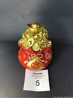 Front view of red and gold piggy bank with Chinese symbols, decorative gold coins, and gold ingot on top next to a Pepsi can for size reference.