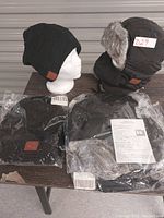 Photo of 2 toques (one displayed on a white mannequin head, one with ear flaps and fur lining) on table with plastic packaging for 2 other similar toques beneath.