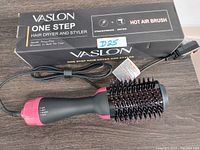 Photo of Vaslon One Step Hair Dryer and Styler Hot Air Brush with its box. The device is black with pink accents, has a brush head with bristles, and a power cord.