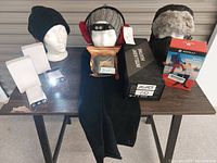 Full view of the winter gear bundle laid out on a dark table, displaying all items with mannequin heads showcasing various toques and earmuffs. Boxes and packaging for heated gloves, socks, and hand warmers visible.