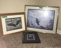 Three framed artworks featuring eagles. One smaller framed eagle head close-up, one medium-sized eagle flying over forest, and one larger artwork of two eagles in a misty forest, displayed on floor.