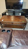 Wooden coffee table with two drawers and decorative carved edges viewed near a brown leather couch and TV stand