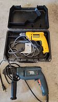 DeWalt drill in open black case and Black & Decker hammer drill with cords coiled on floor.