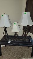 Three wrought iron lamps on a black metal table, each with white fabric bell shades with scalloped edges. Lamps vary in height and base design.