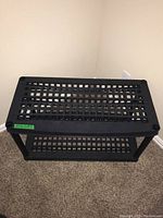 Single black plastic shelving unit against a beige wall on carpet floor, showing grid-patterned shelves and cylindrical legs