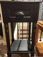 Black wooden side table with drawer showing front view and lower shelf, located indoors near stairs.