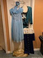 Full view of blue vintage dress with pleated sheer overlay, dark blue dress with white lace collar, original storage bag and accessories hanging on a door.
