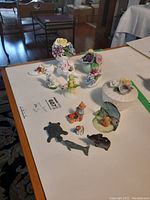 Table display of porcelain floral figurines, animal figurines in stone and resin including turtle, dolphin, small bear, and a lidded trinket dish with floral motif.