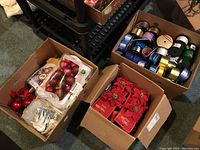 Three cardboard boxes filled with Christmas decorations including bundled red poinsettias, a pack of red balls, white snow baby figurine, bird and sleigh ornaments, faux holly garland, and spools of decorative ribbon in multiple colors.