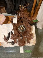 Photo showing full front view of wood cuckoo clock with deer head, crossed rifles, and detailed wood carving. Weights and chains visible on side.