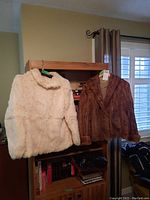 Full view of white rabbit fur jacket and brown fox fur jacket hanging on wooden shelf with window background