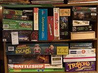 Photo shows top view of board games and puzzles in stacked arrangement. Visible titles include Jurassic Era puzzle, Dinosaur floor puzzle by Melissa & Doug, Game of Thrones 4D Puzzle, Monopoly Junior, Payday, Battleship, Ultimate Operation Escape Room, Lord of the Rings Risk, Mastermind, Trivial Pursuit, and Tribond.