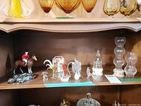 Shelf with crystal glass figurines including swans, birds, glass bell, ceramic horse and rider, model car, and lidded China trinket boxes