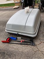Gray Karrite Voyager roof storage box placed on ground with three snow brushes (red, blue, black) with extendable handles in front of it, and a small plastic container on top of the box.