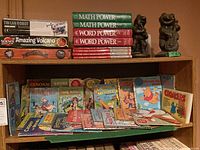 Shelf with two frog figurines, multiple educational book sets (Math Power, Word Power), and science activity kits (Tin Can Robot, Amazing Volcano)