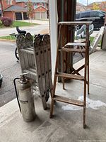 The photo shows an aluminum scaffold ladder folded and standing upright next to a wooden step ladder approximately 4ft tall. A metal fire extinguisher cylinder is placed in front on the floor. The background shows a residential driveway and garage setting.