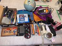 Overview image showing all items arranged on a table; globe, corded phone, calculators, paint palette, writing utensils, baskets, alarm clock, picture frame, and headphones visible.