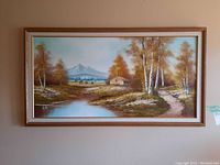 Frontal view of signed framed painting depicting a small house, birch trees, pond, and mountain in the distance in warm autumn colors.