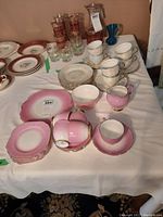 Photo of multiple vintage dishware items arranged on a table, including pink and gold lustreware pieces, white teacups and plates, glass tumblers, and a blue glass pitcher or vase.