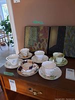 Photo showing all six pairs of vintage teacups and saucers arranged on a wooden surface with decorative wall art in the background.
