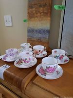 Six vintage floral teacups with saucers arranged on a wooden surface showing varied pink, purple, and blue flower designs with gold trim
