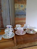 Photo of 6 vintage porcelain teacups and saucers arranged on wooden furniture, each with distinct floral patterns and gold trim.