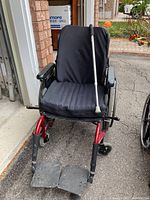 Front view of wheelchair with black fabric cover, red frame, black armrests, and two black footrests. A silver walking cane rests on the seat back.