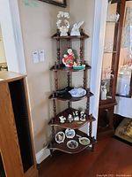 Front side view of wood corner display shelf with five shelves holding various decorative items, placed in a corner beside a large wooden cabinet and near an entryway.