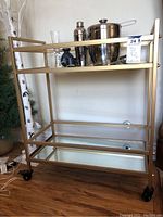 Front view of brass and glass three-tier rolling bar cart with stainless steel barware and drinkware on top shelf.