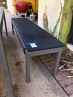 Side view of wood and metal bench with black cushion outside by cactus plants, showing metal legs and weathered cushion.