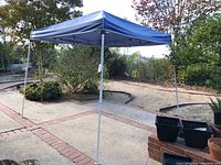 Blue pop-up canopy tent set up on patio, blue fabric roof with visible tears, metal frame appears wobbly.