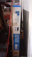 Box of Studio Designs drafting table standing upright in a cluttered garage space with leg of treadmill visible.