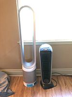 Dyson fan and Lasko heater standing side by side on a wooden floor near a window, showing full front view.