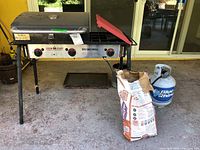 Full front view of the Camp Chef Big Gas Grill showing knobs, lid closed, propane tank and charcoal bag placed in front.
