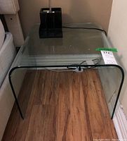 Front left side angle view of curved clear glass end table on wood floor with lamp base on top.