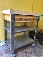Front angle of rolling kitchen island with butcher block wooden top, stainless steel frame, two metal shelves, single drawer, and caster wheels on a concrete floor against yellow wall.