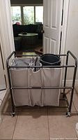 Laundry hamper with three beige fabric bags and a black plastic bucket on a metal frame with wheels.