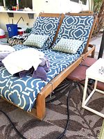 Front angle view of the wooden lounge chair with patterned cushions and pillows, bedding folded on top, placed on outdoor rug near pool area.