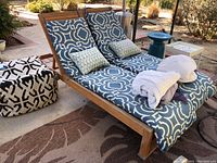 Lounge chair with matched blue and white patterned cushions, two smaller pillows, and rolled white bedding on top, sitting on patio with rug and nearby decor