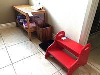 Wooden spa bench with lower shelf, red step stool, brown waste basket, and black bathroom scale arranged on tiled floor near wall.