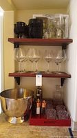 Shelves showing six clear stemmed wine glasses on the middle shelf, three black textured stemmed goblets, clear water jug on the top shelf, silver-plated ice bucket on the floor, and red serving tray holding glass tumblers and salt and pepper shakers.