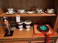 Photo of four tea cups with matching saucers arranged on a wooden shelf along with two white vases, a decorative doll, and a carved red wooden stand with green jade-like centerpiece.