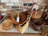 Overall view of multiple wicker baskets including round baskets, a rectangular plate holder, and a large urn-shaped basket with lid and ribbons. Decorative ceramic bowl with lattice design and candle holder inside at right.