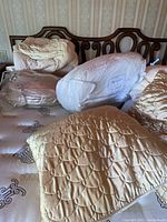 Photo showing various king size bedding items including quilted down blankets, mattress pads and packaged bedding items on a bed with dark wood headboard.