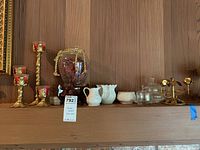 View of the lot on wooden mantle showing the candleholders, red glass vase, white ceramic pieces, and glass boxes.
