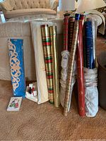 Photo showing multiple rolls of Christmas wrapping paper in various colors and patterns along with Christmas greeting cards and additional roll on couch.