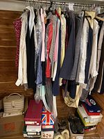 Photo of hanging women's clothes on rack showing dresses, blouses, jackets in various colors and fabrics, some protected by plastic sleeves. Below are shoe boxes and a beige handbag.
