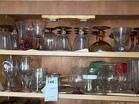 Photo of wooden shelf with two shelves holding various clear and colored glassware and stemware including goblets, wine glasses, bowls, and tumblers.