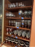 Wood cabinet shelves showing an assortment of clear glass stemware, including wine glasses, goblets, and cocktail glasses on four shelves.