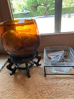 Amber glass bowl with three amber glass balls inside on wooden stand next to clear square glass planter with rocks, on carpet near window